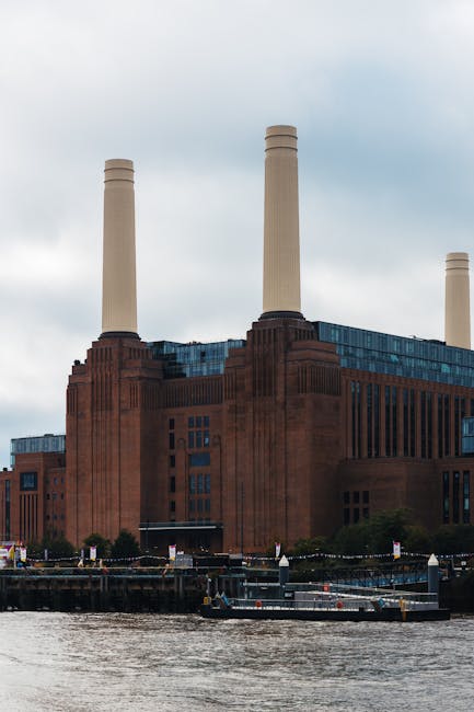 The image shows the iconic Battersea Power Station with its distinctive brick facade and four tall white chimneys set against a cloudy sky. In the foreground, part of the river Thames is visible, along with a dock area where a flatbed trolley is loaded with several cardboard boxes and wrapped furniture items, likely in the process of a home relocation. Nearby, a large white van with open doors is parked on a paved area, facilitating furniture transport and packing materials for moving services. The scene captures a professional removal operation by Removal Companies Battersea, emphasizing the loading process of household belongings, with orange straps and protective blankets visible on the furniture items to prevent damage during transport. The environment is urban, with trees and a structured dock visible, illustrating an organized moving setup within the property’s exterior, supporting efficient and secure furniture transport from the residence to the new destination.