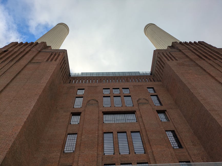 A low-angle photograph of the iconic Battersea Power Station, showing its red brick facade with multiple rectangular and arched windows. The image captures the two tall, cylindrical chimneys extending upward into a cloudy sky, emphasizing the building's industrial architecture. The perspective highlights the scale of the structure, often involved in home relocation and furniture transport projects. Inside or near the power station, there is no visible furniture or moving equipment, but the historical building's exterior suggests preparation for a house removal or relocation service. The setting implies an urban environment with the building's robust, utilitarian design and substantial height, which may be relevant to relocation companies like Removal Companies Battersea when planning logistics for moving furniture or household items to or from the site.
