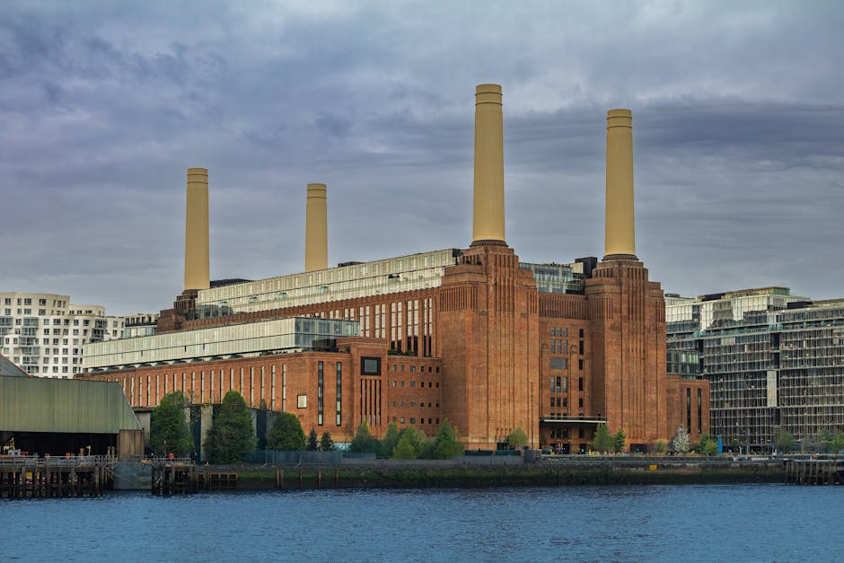 A large industrial building with a brown brick exterior and four tall, cylindrical smokestacks extending upward. The structure is situated beside a body of water, with a green, metal-roofed shed and a wooden dock visible in the foreground. Residential and commercial buildings surround the site, and the sky overhead is cloudy. This scene may depict an energy plant or factory located near a waterfront, with the setting suitable for house removals and moving services such as furniture transport or home relocation activities supported by Removal Companies Battersea, as seen in the context of their services on the Clapham Junction to Battersea removals solutions page.