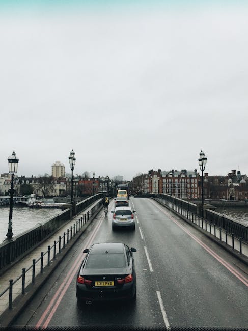 The image shows a wide view of a bridge over a river, with a line of moving vehicles, including a dark grey car in the foreground and a white van further ahead, heading towards the cityscape. The bridge is equipped with decorative black lampposts on both sides, and the road surface appears damp, suggesting recent rain. On either side of the bridge, red brick and stone buildings line the riverbank, some with chimneys and flat roofs, indicating an urban area. The sky is overcast with grey clouds, creating a subdued lighting environment. This setting resembles a typical urban transportation route used during house removals or furniture transport when moving between locations such as Clapham Junction and Battersea, with the scene capturing the typical traffic flow and cityscape in the background. The image subtly reflects aspects of moving logistics and urban moving services provided by companies like Removal Companies Battersea.