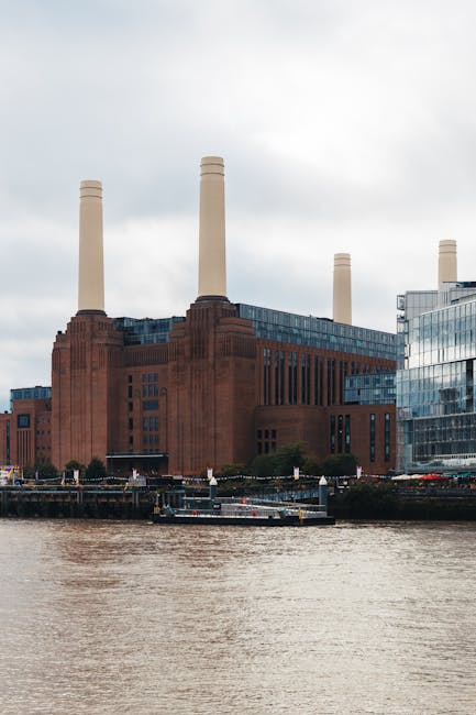 A large industrial building with a brown brick exterior and four tall, cylindrical smokestacks extending upward. The structure is situated beside a body of water, with a green, metal-roofed shed and a wooden dock visible in the foreground. Residential and commercial buildings surround the site, and the sky overhead is cloudy. This scene may depict an energy plant or factory located near a waterfront, with the setting suitable for house removals and moving services such as furniture transport or home relocation activities supported by Removal Companies Battersea, as seen in the context of their services on the Clapham Junction to Battersea removals solutions page.
