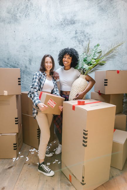 Two women are standing inside a property surrounded by numerous packed cardboard moving boxes, some sealed with red packing tape. One woman, dressed in a plaid shirt and beige trousers, holds a medium-sized box with a red 'fragile' sticker, while the other, wearing a white top and patterned trousers, carries a large decorative vase with green palm leaves. The background features a textured, light grey wall, and the floor is wooden. The scene depicts a home relocation process where the women are preparing for a move, with the boxes arranged around them indicating packing and furniture transport activities. Removal Companies Battersea is involved in this packing and moving process, ensuring smooth logistics for a house removal or house moving service.
