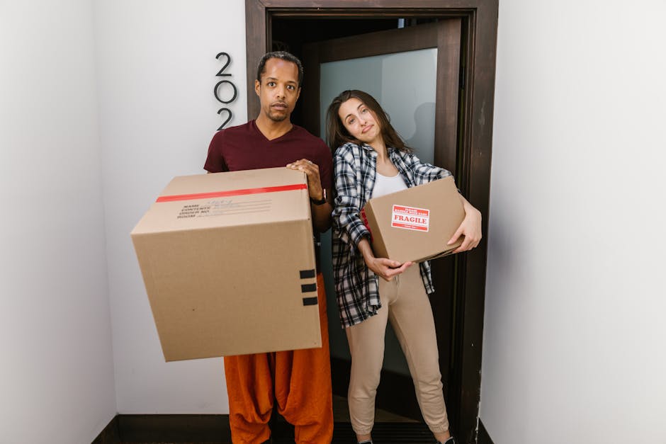 Two movers from Removal Companies Battersea are standing inside a residential doorway, each holding a cardboard box for house relocation. The male mover on the left is wearing an orange outfit and a maroon t-shirt, carrying a large, sealed cardboard box with a shipping label visible. The female mover on the right is dressed in beige pants and a white top with a plaid shirt, holding a smaller box marked with a red 'Fragile' sticker. The background shows a white wall with house number '202' mounted vertically beside the door frame, and the flooring is dark-colored. The door, positioned behind the movers, is slightly open, revealing the interior of the property. The scene captures the early stages of packing and furniture transport during home moving, with lightweight, packed boxes ready for loading onto a van for a smooth move, illustrating the physical logistics involved in house removals often managed by professional services like Removal Companies Battersea.