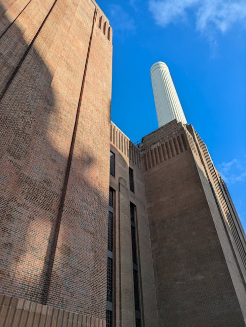 A low-angle view of a tall brick building with narrow vertical windows, partially shaded by shadows cast by nearby structures, under a clear blue sky. The building's exterior features a blend of brick